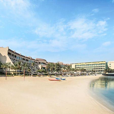 알 라하 비치 호텔 아부다비 외부 사진 The photo depicts a serene beach scene with a clear blue sky. In the foreground, there is a sandy beach that gently slopes down to calm water. On the left side, several palm trees provide a tropical atmosphere. In the background, there are two large 