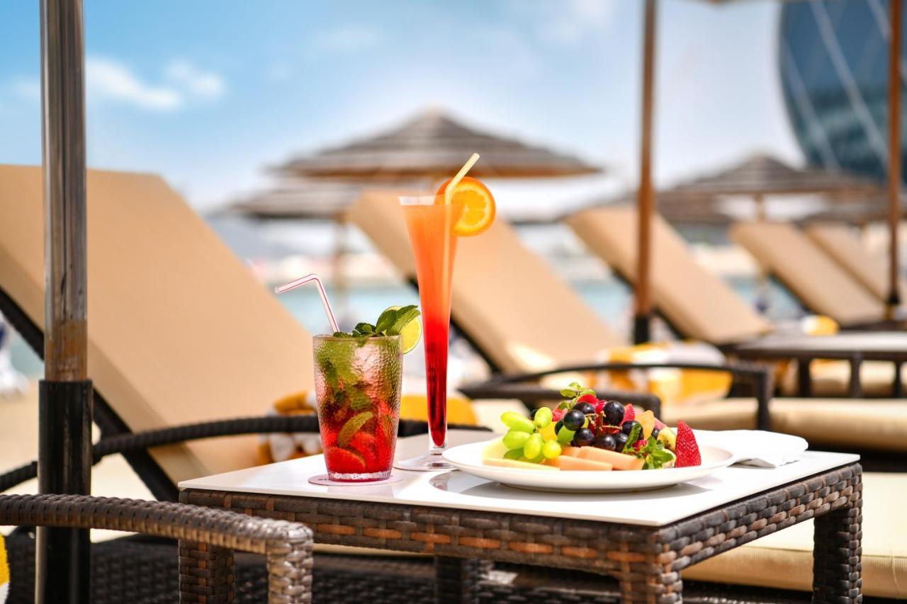 알 라하 비치 호텔 아부다비 외부 사진 The photo shows a table set outdoors, featuring two colorful cocktails and a plate of food. One cocktail is a vibrant red drink garnished with mint, while the other is a layered orange beverage with an orange slice on the rim. The plate of food inclu