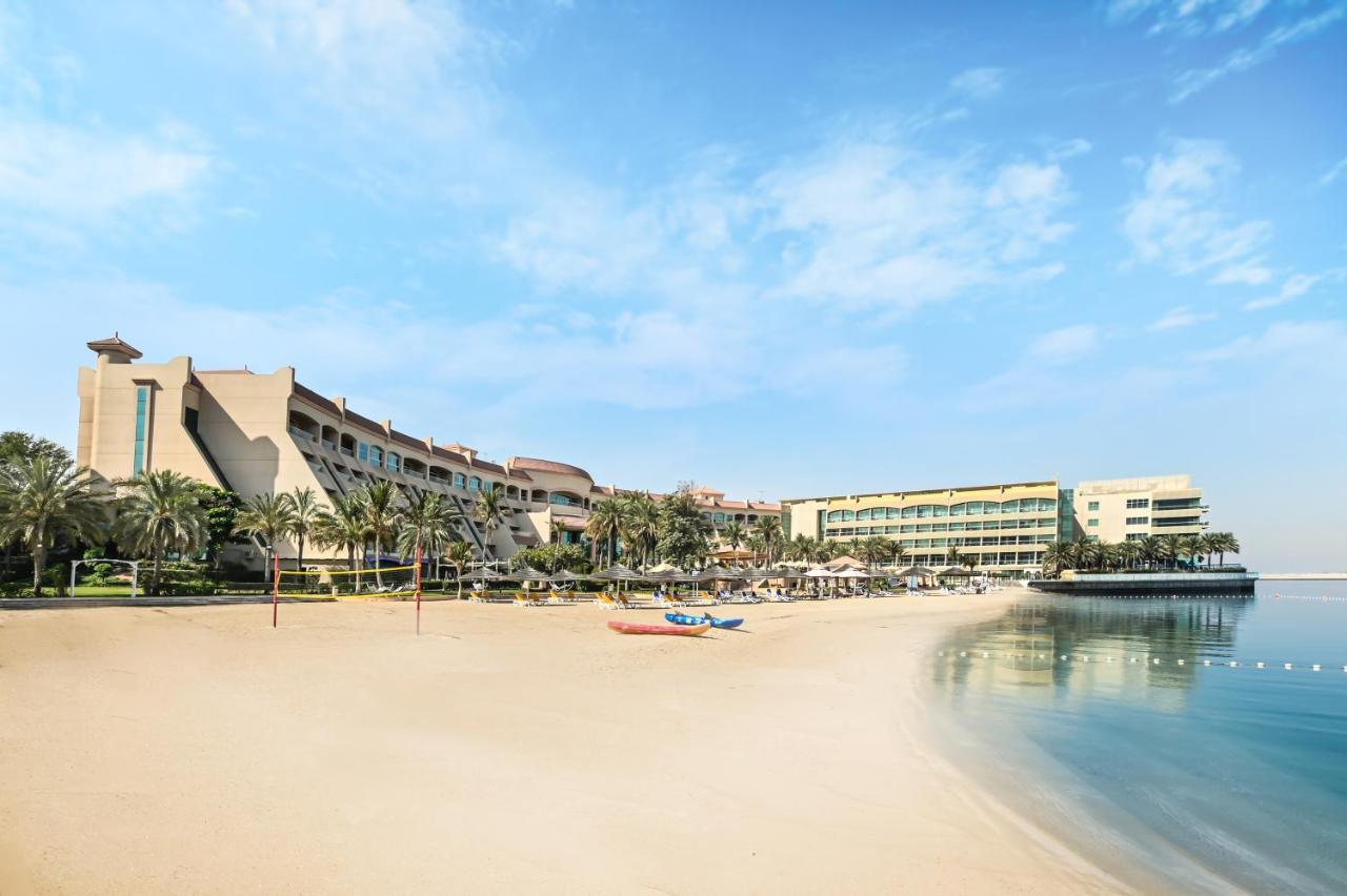 알 라하 비치 호텔 아부다비 외부 사진 The photo depicts a serene beach scene with a clear blue sky. In the foreground, there is a sandy beach that gently slopes down to calm water. On the left side, several palm trees provide a tropical atmosphere. In the background, there are two large 