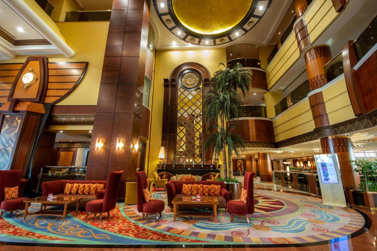 알 라하 비치 호텔 아부다비 외부 사진 The photo shows an elegant lobby interior of a hotel. The space features tall ceilings with decorative lighting and large windows, allowing for natural light. There are several plush seating arrangements, including red armchairs and wooden tables, cr