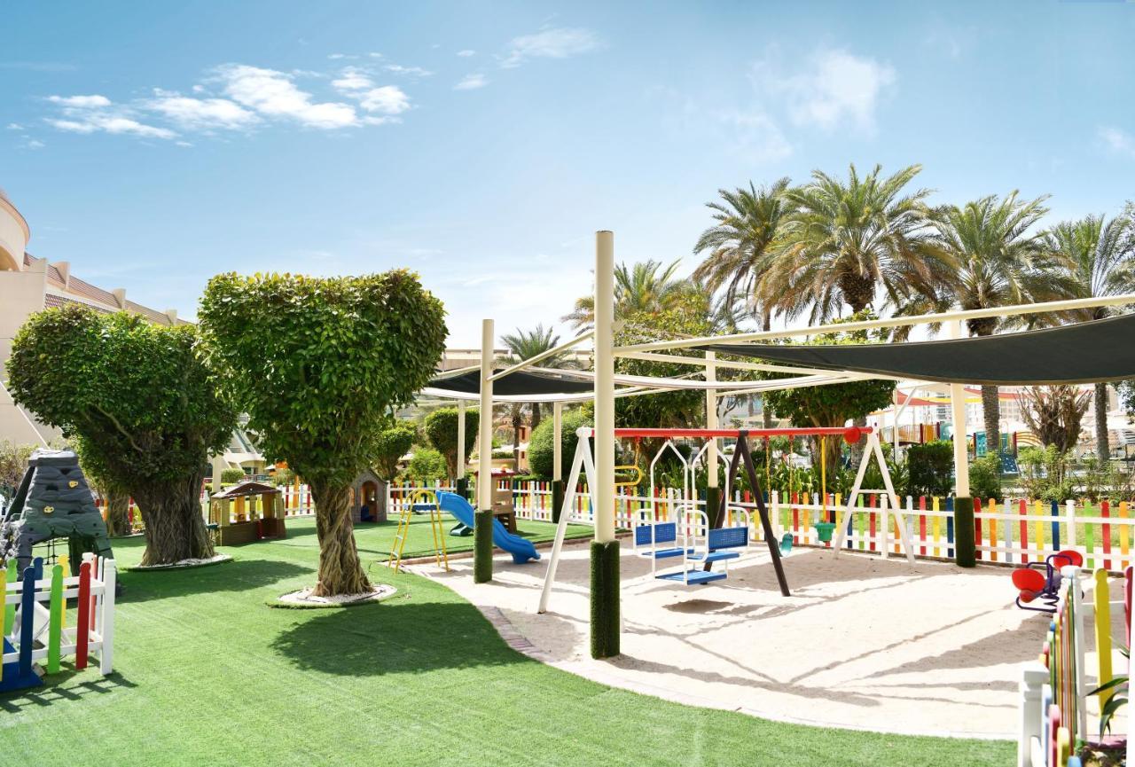 알 라하 비치 호텔 아부다비 외부 사진 The photo shows a brightly lit outdoor playground surrounded by palm trees. The playground features a variety of play equipment, including swings, a slide, and a climbing structure. The ground is covered in lush green artificial grass, providing a so
