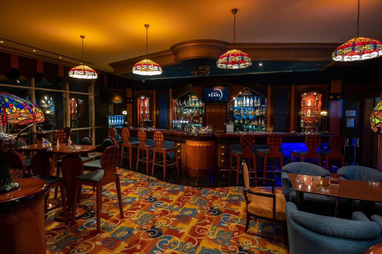 알 라하 비치 호텔 아부다비 외부 사진 The photo shows a cozy bar interior, featuring a well-stocked bar area with various bottles displayed on shelves. The bar is illuminated by colorful stained glass light fixtures, creating a warm and inviting atmosphere. The floor is covered with a vi