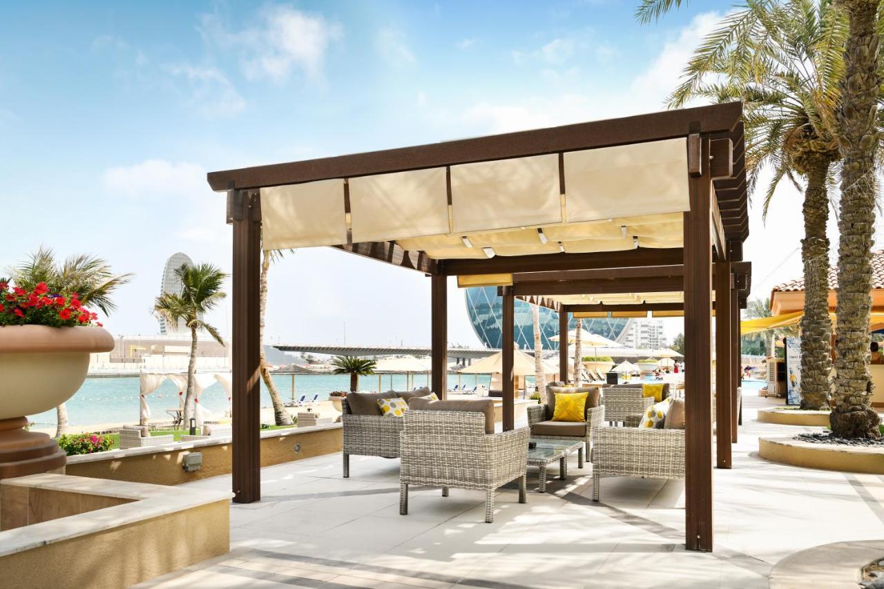 알 라하 비치 호텔 아부다비 외부 사진 The photo shows a stylish outdoor seating area by a beach. There is a wooden pergola covered with a light fabric shade, providing shelter to a set of comfortable wicker chairs and tables. The area is adorned with palm trees, and in the background, th