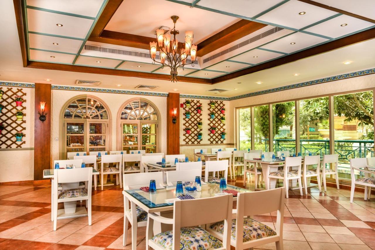 알 라하 비치 호텔 아부다비 외부 사진 The photo shows the interior of a restaurant or dining area. The space has a warm and inviting atmosphere, featuring a mix of decorative elements. 

- **Seating**: There are several white wooden tables and chairs arranged throughout the area, some wi