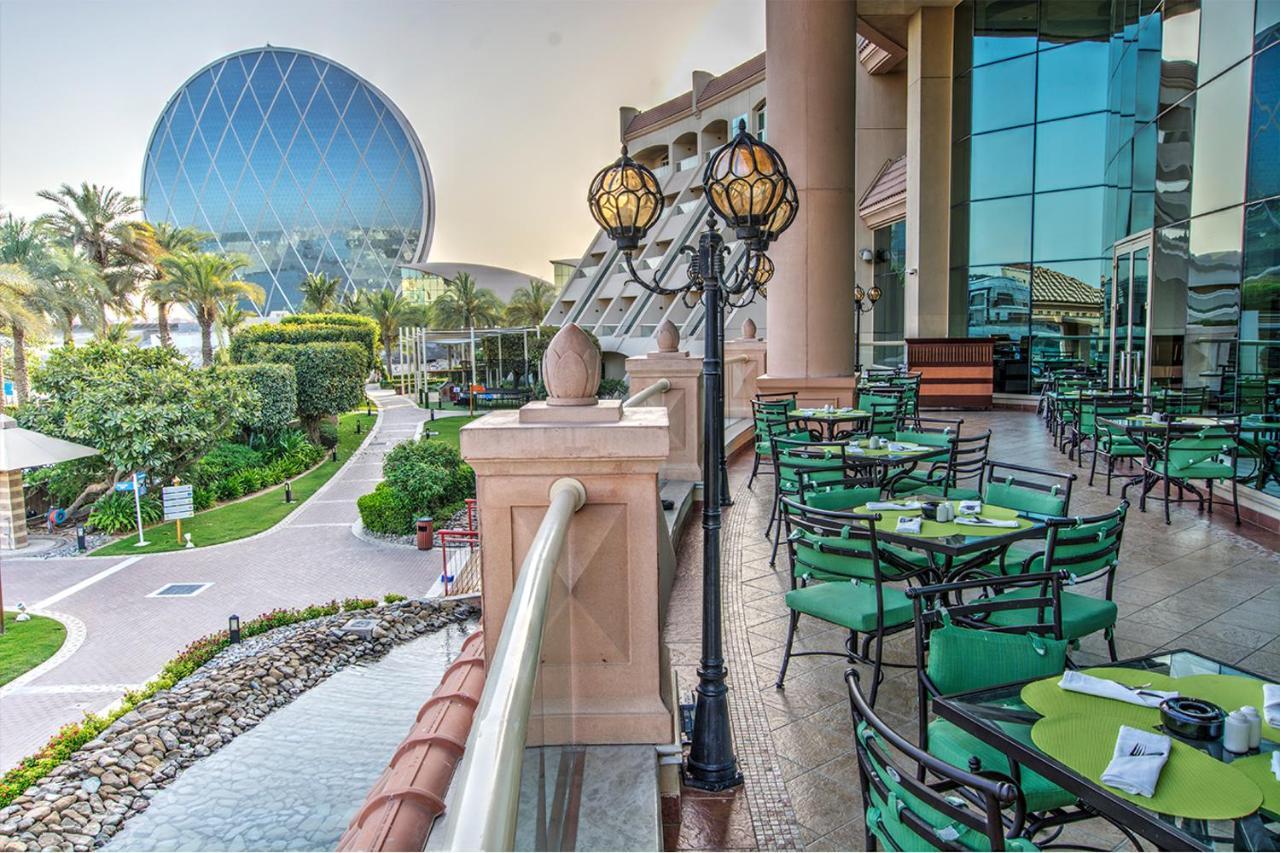 알 라하 비치 호텔 아부다비 외부 사진 The photo depicts an outdoor dining area with green tables and chairs arranged on a patio. In the background, there is a large, distinctive building with a striking blue dome, surrounded by well-maintained landscaping, including trees and plants. The