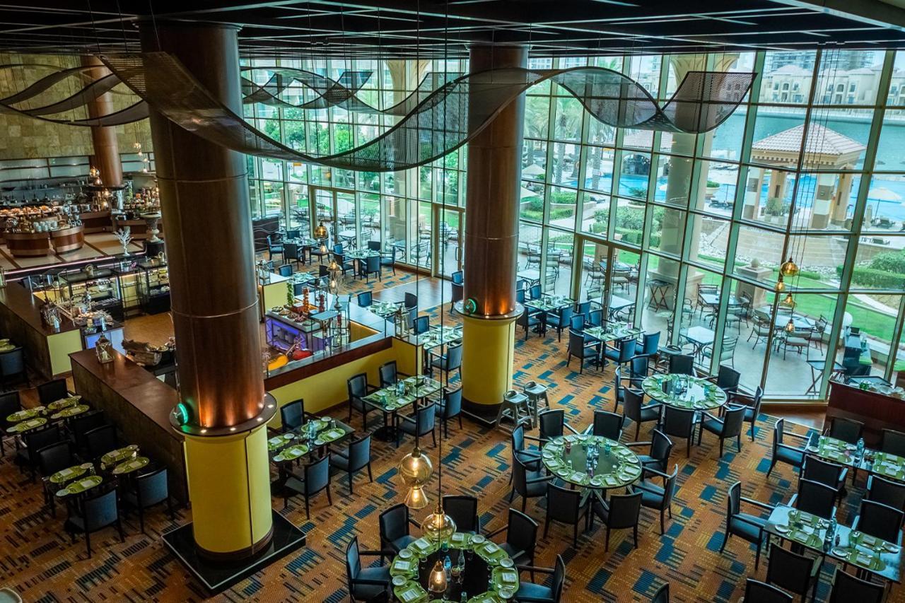 알 라하 비치 호텔 아부다비 외부 사진 The photo shows the interior of a restaurant with a modern design. It features large windows allowing natural light to flood the space, creating an inviting atmosphere. The dining area is furnished with round tables covered with green tablecloths and