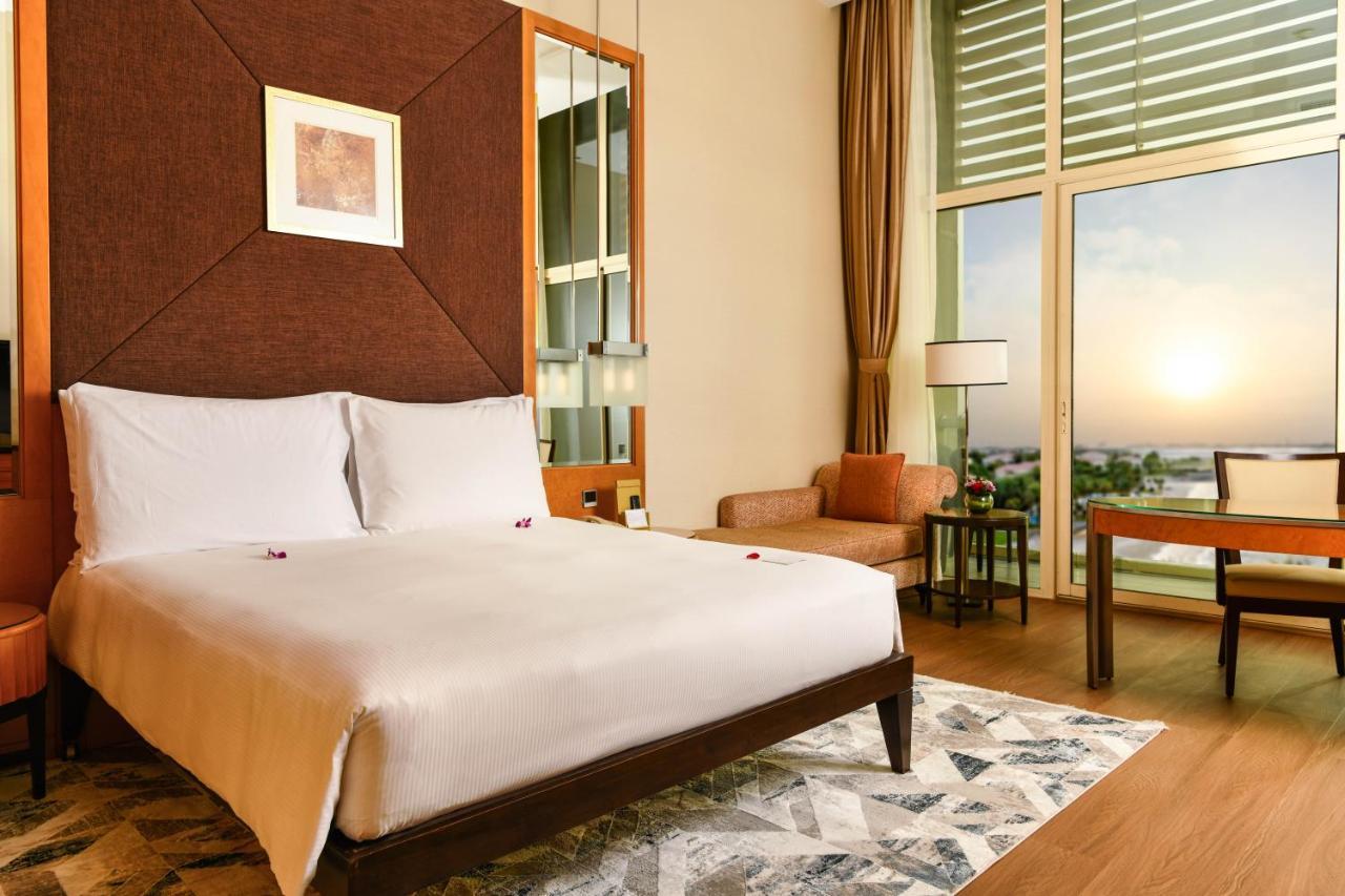 알 라하 비치 호텔 아부다비 외부 사진 The photo shows a stylish hotel room featuring a large bed with white bedding, positioned against a textured wall. There’s a small seating area with a chair and a table, and a window with curtains that allows natural light to fill the space. The room