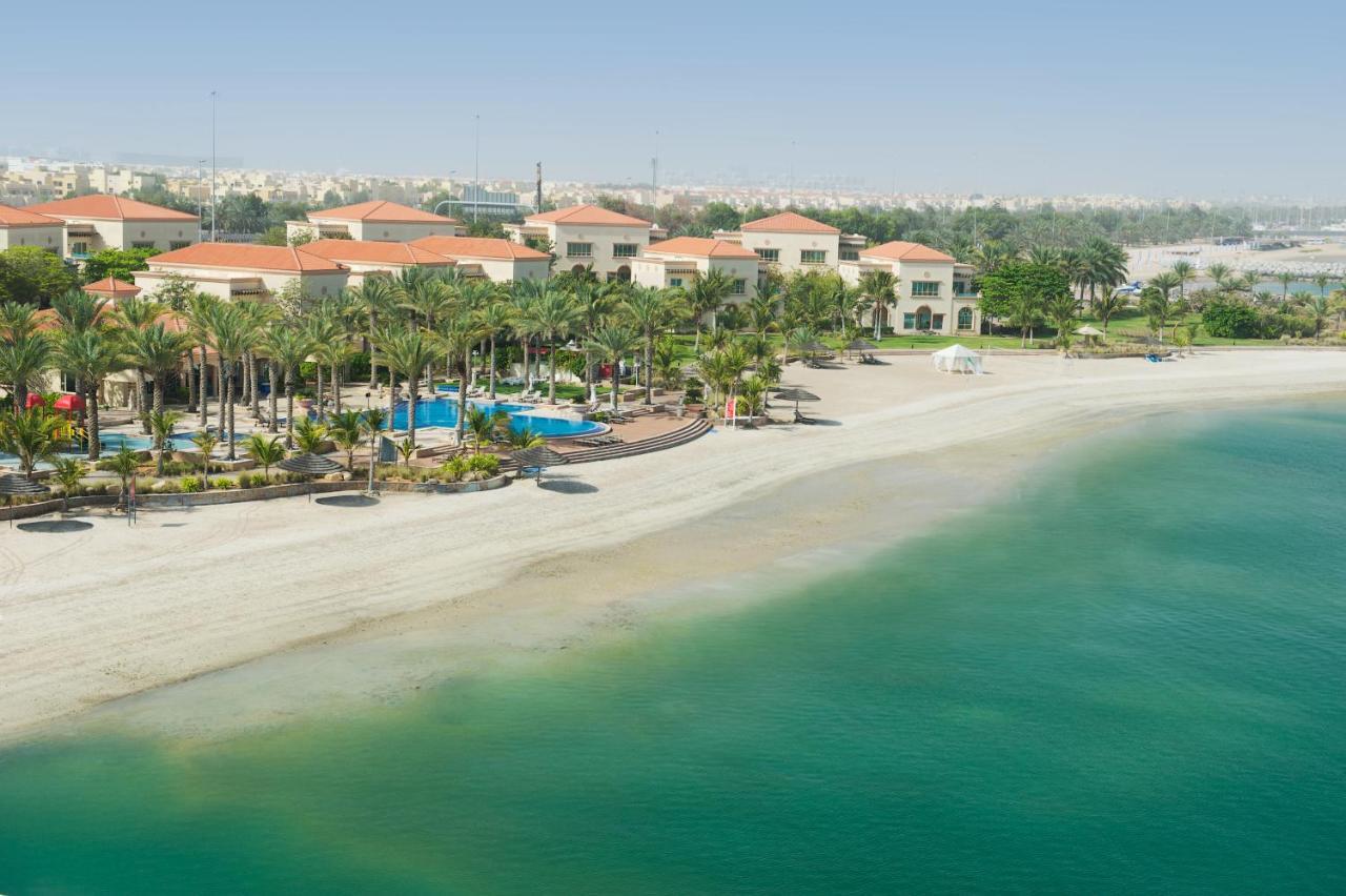 알 라하 비치 호텔 아부다비 외부 사진 The photo depicts a serene beach scene with a clear turquoise sea and a sandy shoreline. In the background, there are several buildings featuring a Mediterranean architectural style with red-tiled roofs. The area is adorned with palm trees and outdoo