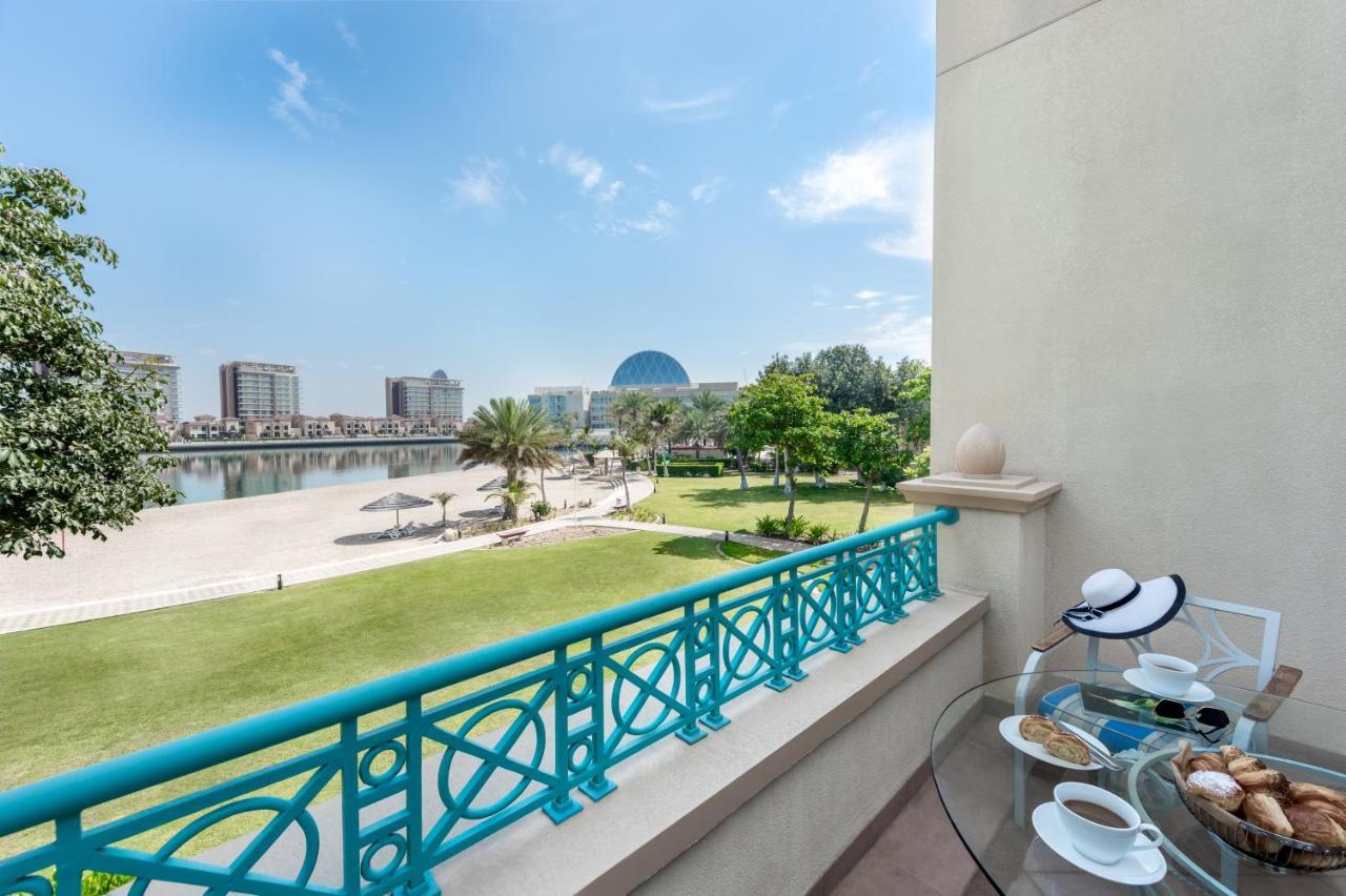 알 라하 비치 호텔 아부다비 외부 사진 The photo shows a balcony view overlooking a scenic landscape. In the foreground, there is a glass table set with snacks or pastries and two cups. Beyond the balcony railing, there is a well-maintained green area with palm trees and a pathway. The ba