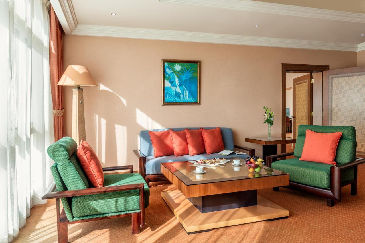 알 라하 비치 호텔 아부다비 외부 사진 The photo shows a cozy living room with a warm ambiance. There are two sofas: one in green with red cushions and another in blue with red cushions, arranged around a wooden coffee table. On the table, there are some fruits and a small decorative item
