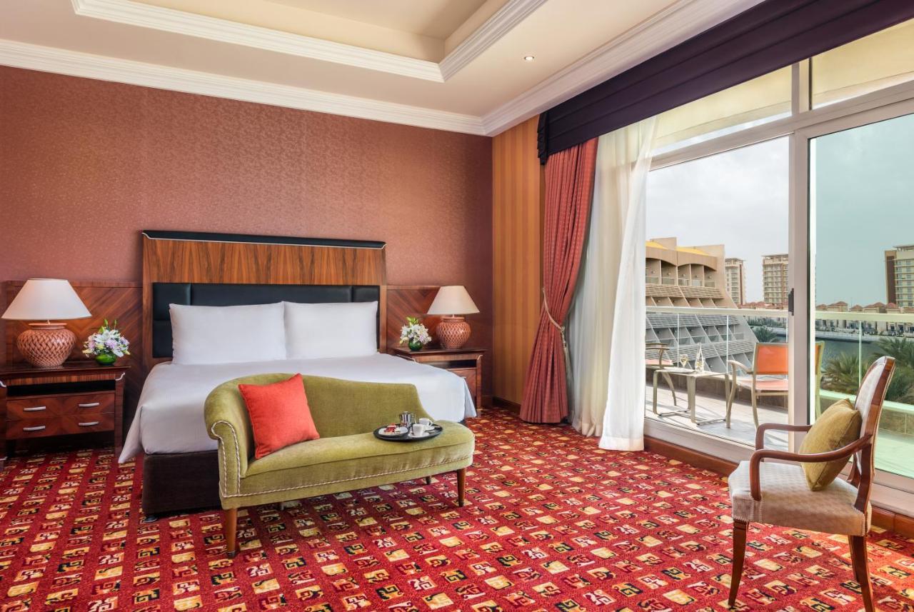 알 라하 비치 호텔 아부다비 외부 사진 The photo shows a hotel room with an elegant and modern design. It features a large bed with white linens and a dark wooden headboard. On either side of the bed are matching bedside lamps. There is a green armchair with a red pillow positioned near t