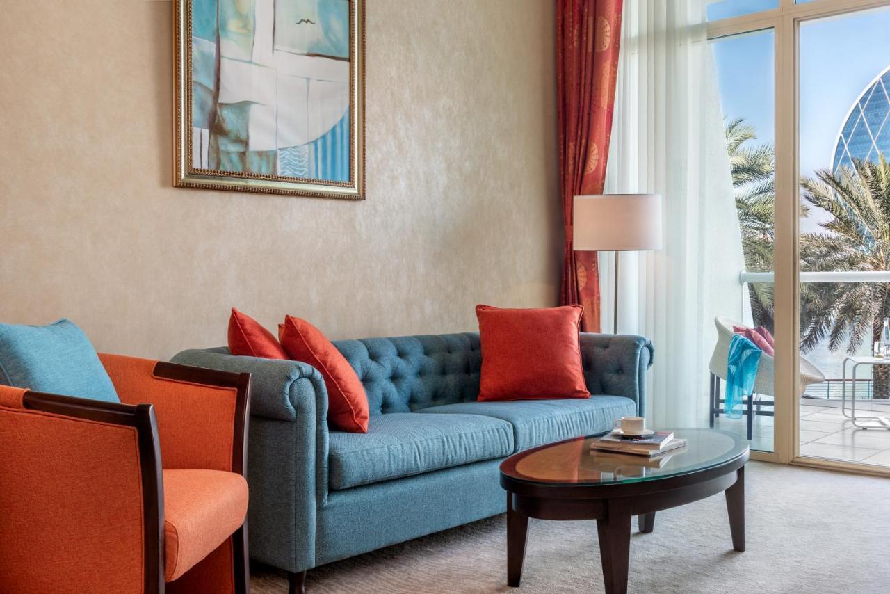 알 라하 비치 호텔 아부다비 외부 사진 The photo shows a well-decorated living room. There is a plush blue couch with orange accent pillows and a cozy orange armchair nearby. A round coffee table sits in front of the couch, and above it, there is a colorful painting hanging on the wall. L