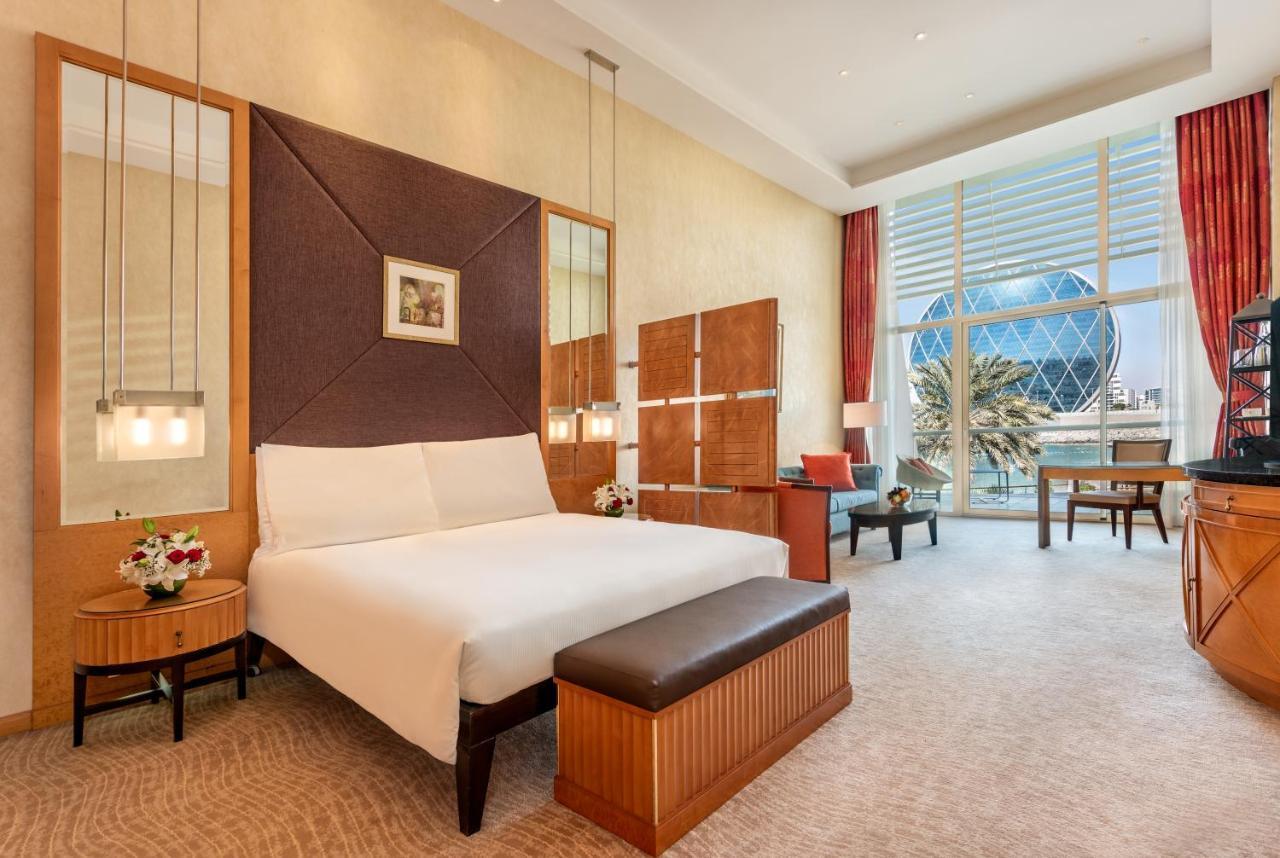 알 라하 비치 호텔 아부다비 외부 사진 The photo shows a modern hotel room featuring a neatly made bed with a white comforter, situated against a stylish headboard with a geometric design. There are bedside tables on either side of the bed, each with a small flower arrangement. A comforta