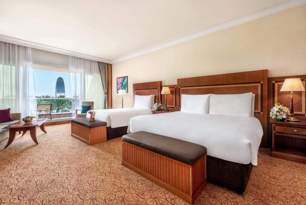 알 라하 비치 호텔 아부다비 외부 사진 The photo shows a spacious hotel room featuring two large beds with white linens and dark brown upholstered headboards. There are matching benches at the foot of each bed. The room is decorated with a warm color palette and has a carpeted floor. To o
