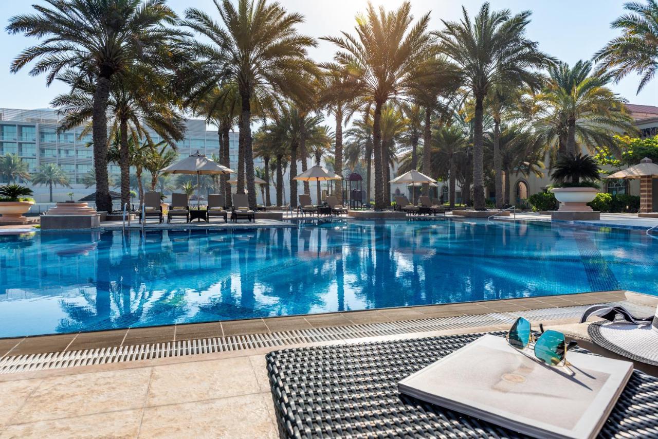 알 라하 비치 호텔 아부다비 외부 사진 The photo shows a vibrant swimming pool surrounded by palm trees under a clear sky. The pool's water is sparkling in the sunlight, creating a serene and inviting atmosphere. Lounge chairs are placed around the pool, some of which are shaded by umbrel