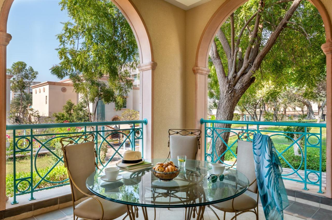 알 라하 비치 호텔 아부다비 외부 사진 The photo shows a serene outdoor balcony setting. There is a circular glass table with a bowl of fruit or snacks in the center, alongside a glass and a small plate. Surrounding the table are four elegant chairs. The railing is turquoise, framing a vi