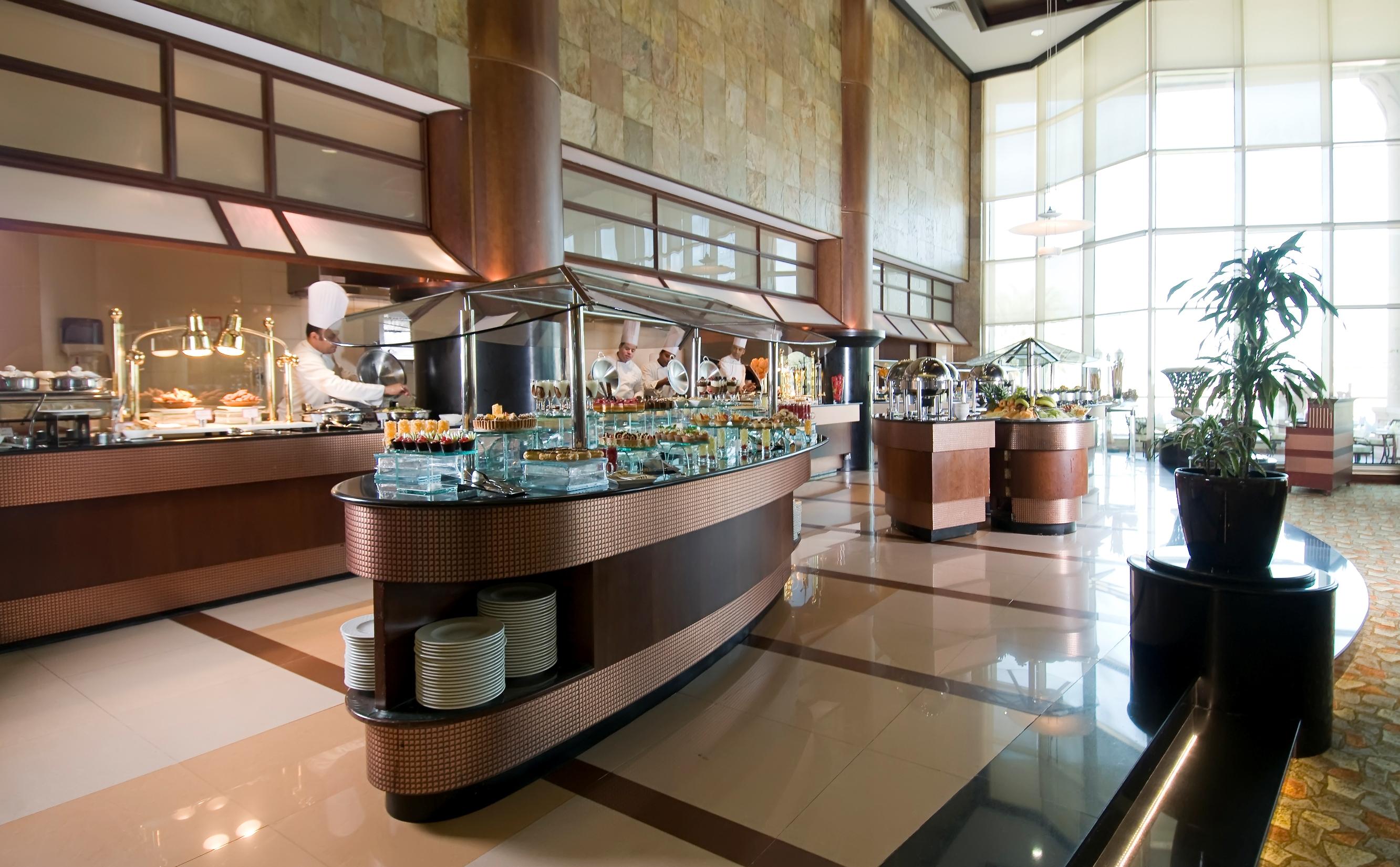 알 라하 비치 호텔 아부다비 외부 사진 The photo shows a spacious buffet area in a restaurant or hotel. There are multiple food stations with glass displays showcasing a variety of dishes. Chefs can be seen in the background, preparing food. The decor features large windows allowing natur