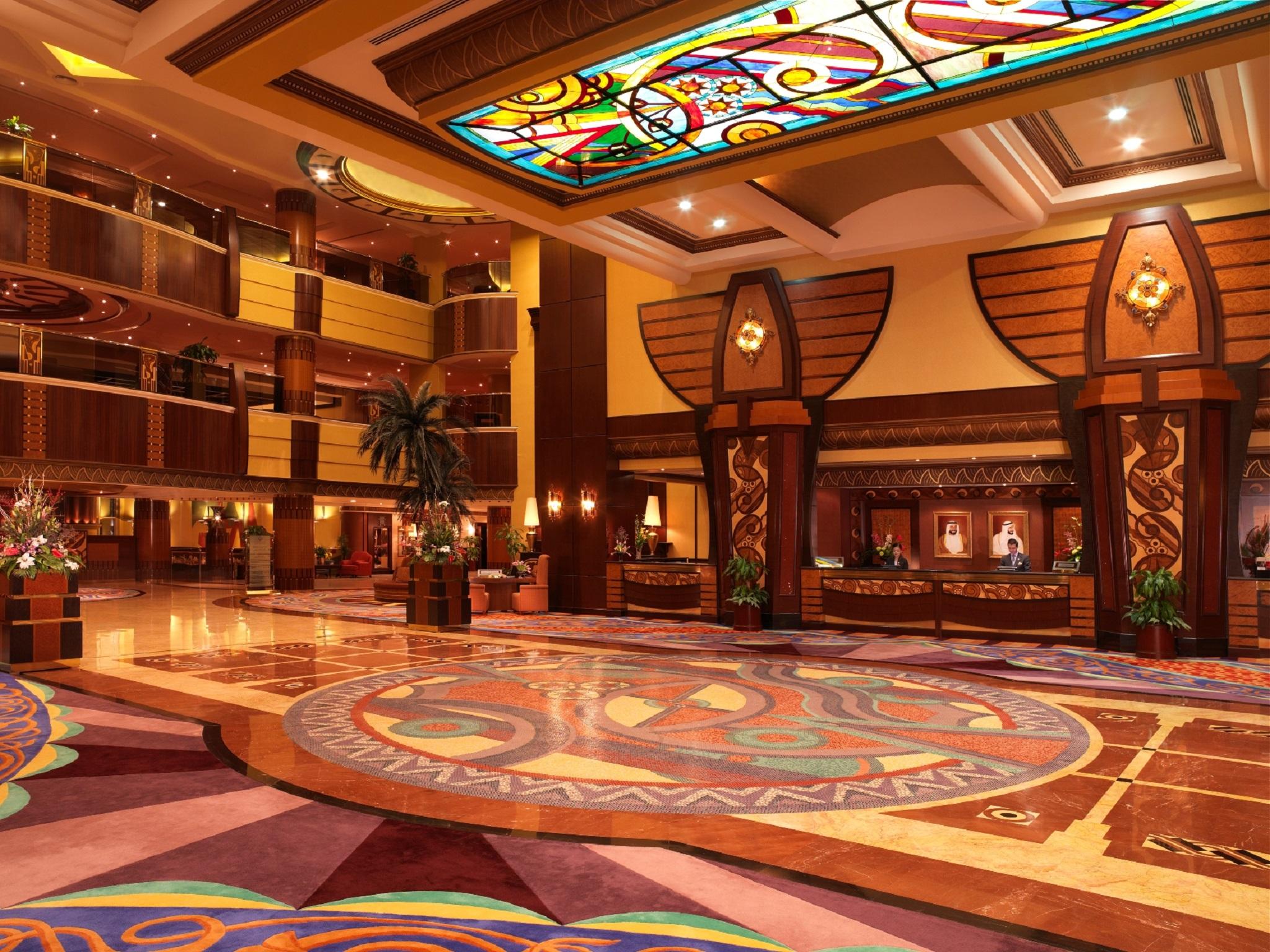 알 라하 비치 호텔 아부다비 내부 사진 The photo depicts an elegantly designed hotel lobby. It features a grand ceiling with intricate stained glass and decorative patterns. The floor is adorned with colorful, ornate designs, including circular patterns. The lobby is spacious, with wooden
