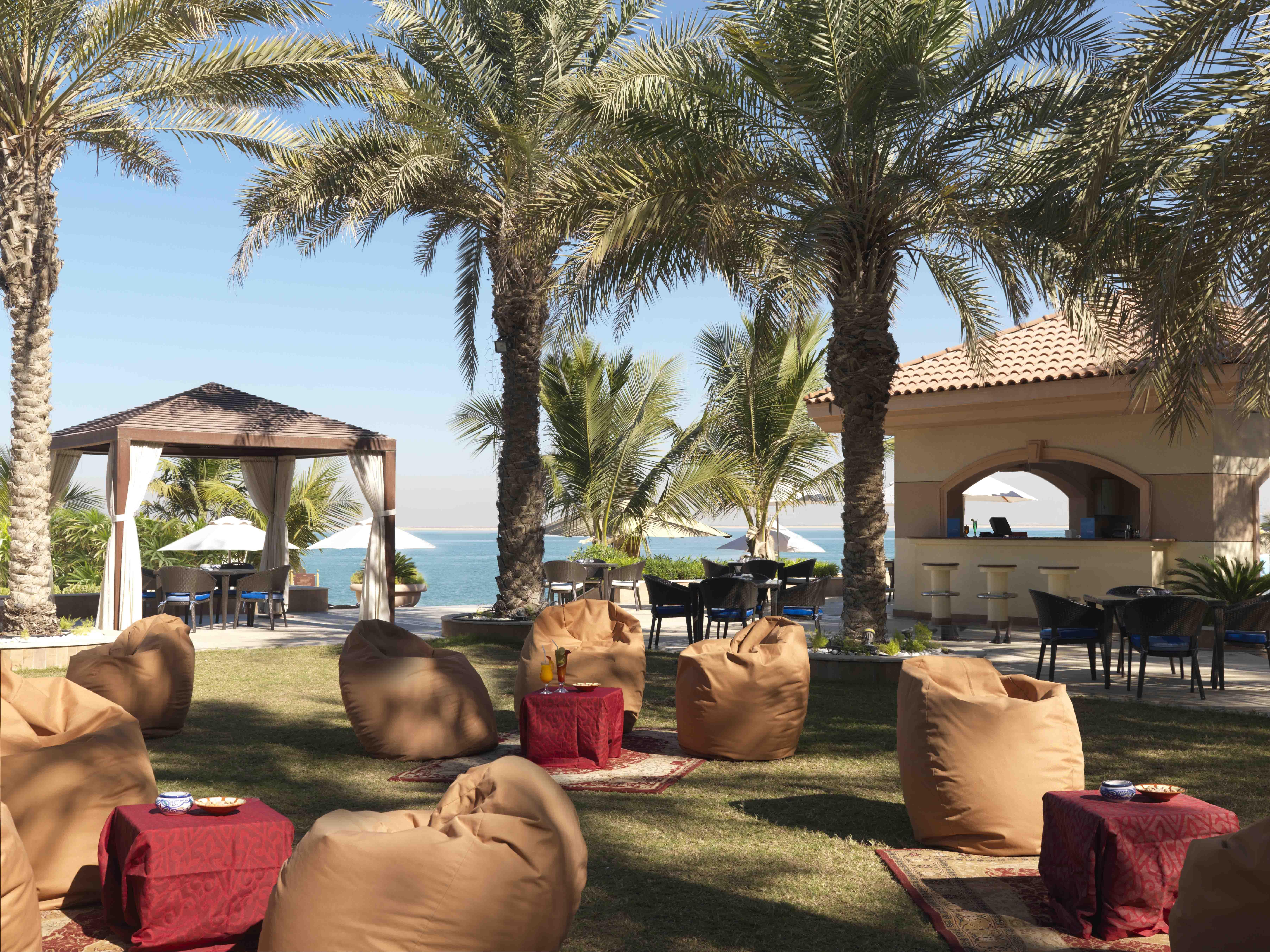 알 라하 비치 호텔 아부다비 레스토랑 사진 The photo depicts a beautiful outdoor setting with several elements of relaxation and leisure. In the foreground, there are large, comfortable bean bag chairs arranged on a grassy area, accompanied by low tables. Surrounding this seating area are tal