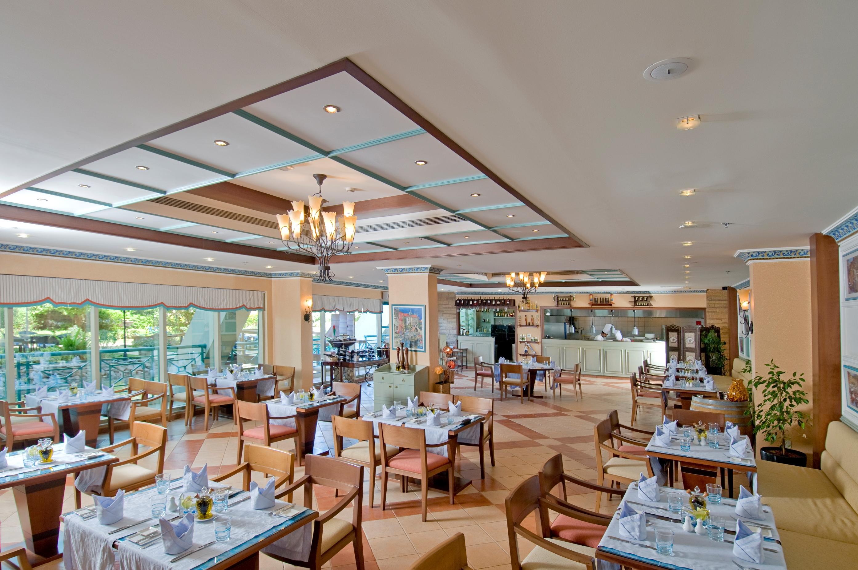 알 라하 비치 호텔 아부다비 레스토랑 사진 The photo shows the interior of a restaurant. It features a spacious dining area with multiple tables neatly arranged, each set with tableware and cloth napkins. The decor includes a decorative ceiling with lighting fixtures, and large windows that p