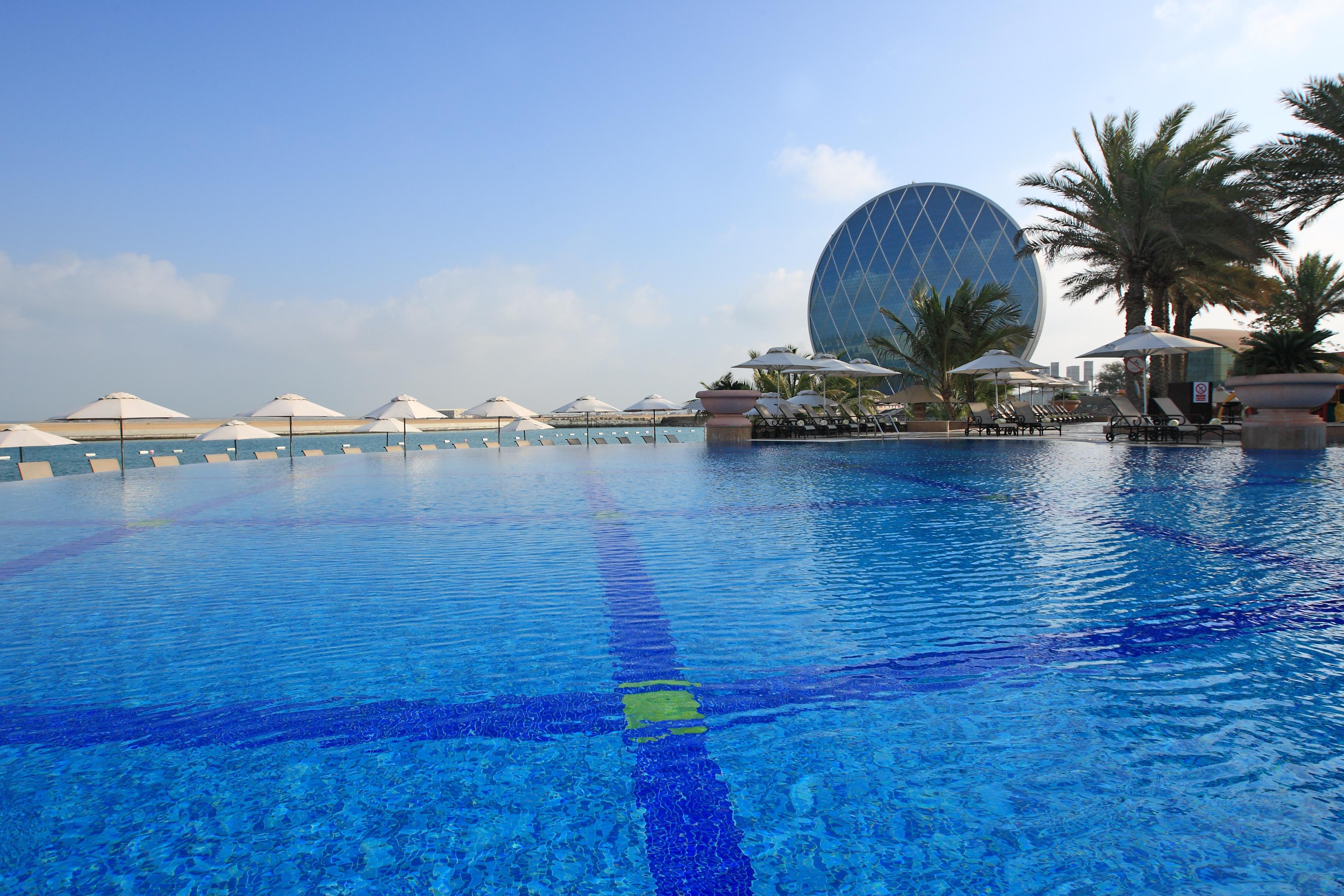 알 라하 비치 호텔 아부다비 외부 사진 The photo features a large, serene swimming pool with clear, blue water reflecting the sky. Surrounding the pool are several lounge chairs and umbrellas, suggesting a relaxed, resort-like atmosphere. In the background, a distinctive building with a r