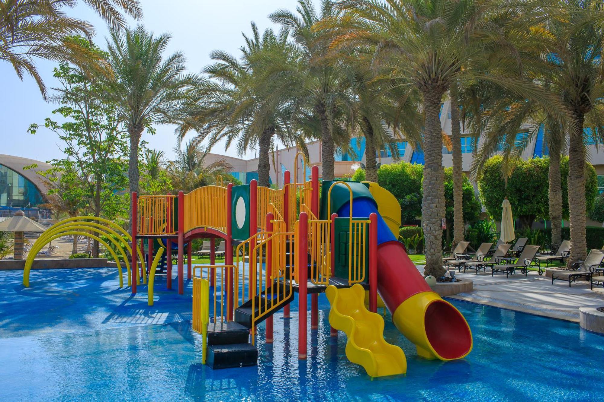 알 라하 비치 호텔 아부다비 외부 사진 The photo shows a colorful water play area located in a pool setting. It features a variety of play structures, including slides in multiple colors, such as yellow, red, and blue. Surrounding the play structure are palm trees, creating a tropical atm
