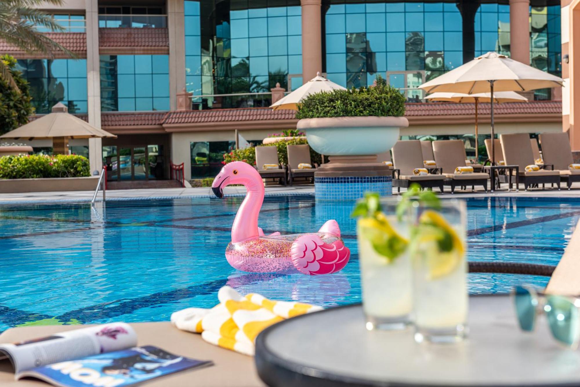 알 라하 비치 호텔 아부다비 외부 사진 The photo depicts a bright and inviting outdoor swimming pool surrounded by a luxurious setting. In the pool, there is a large pink flamingo float, adding a fun and whimsical touch. To the side, a table is set up with two refreshing drinks, likely co