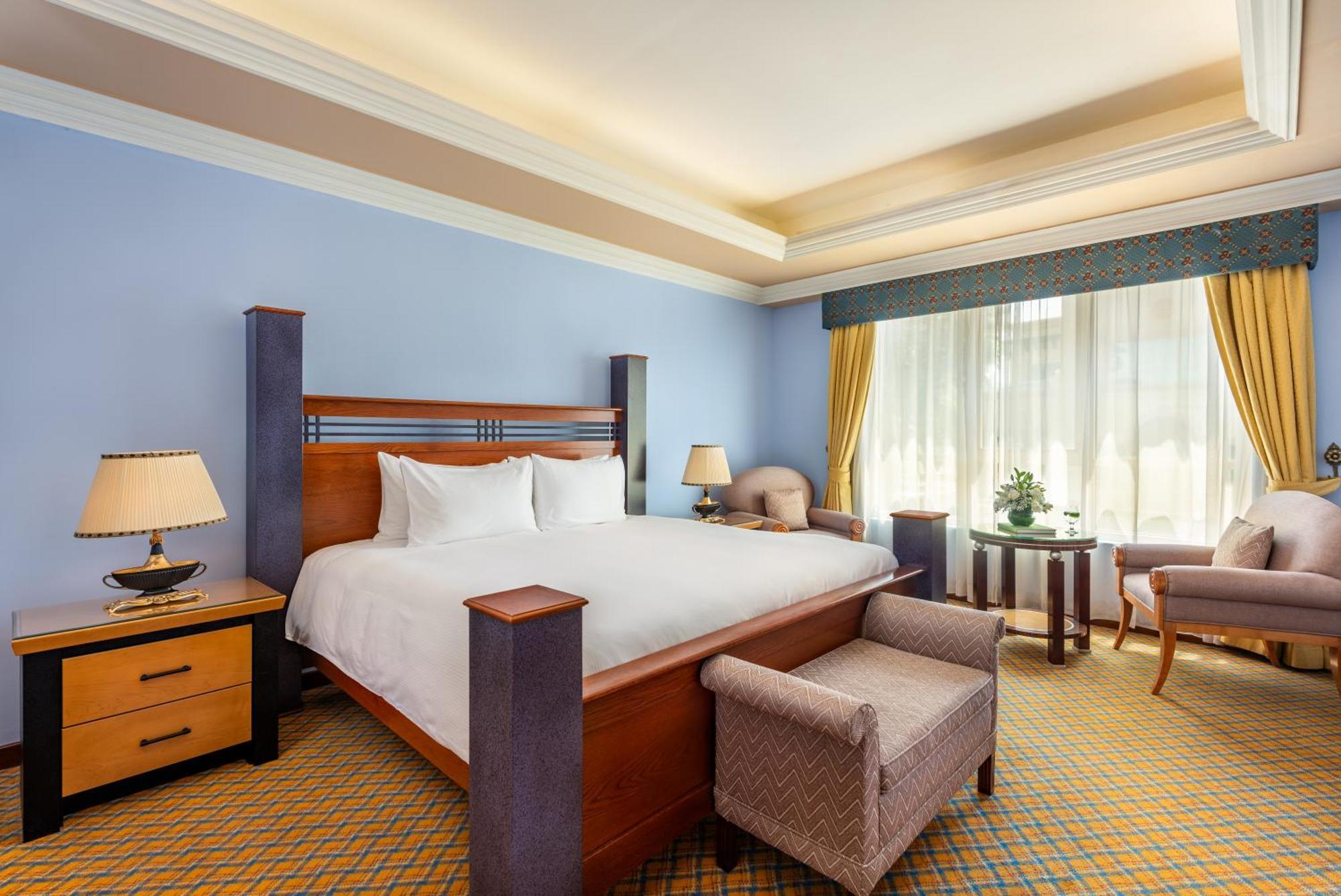 알 라하 비치 호텔 아부다비 외부 사진 The photo shows a well-decorated hotel room. It features a large bed with a wooden frame, neatly made with white bedding. On either side of the bed are bedside tables with lamps. There is a comfortable chair next to a window that has sheer curtains a