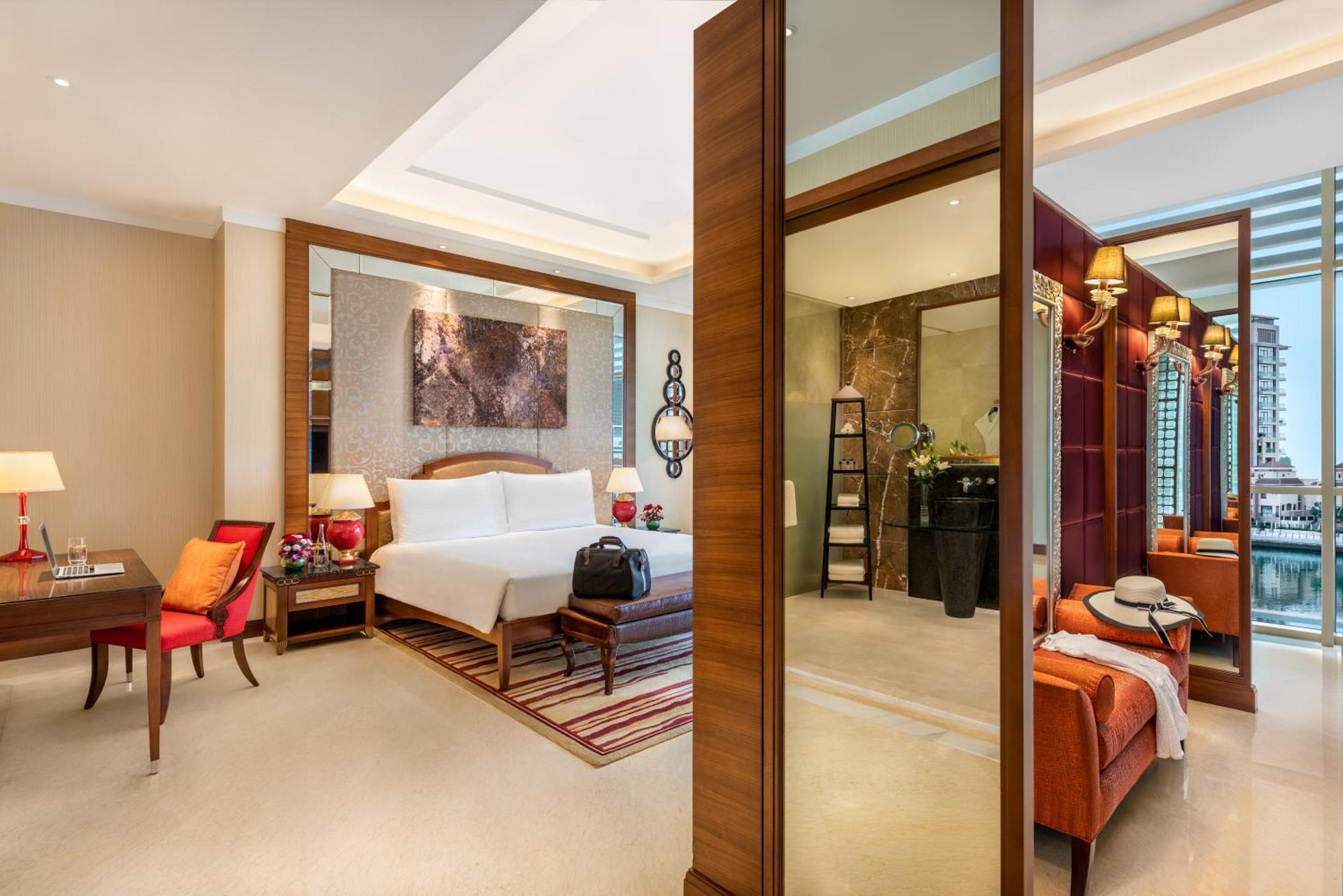 알 라하 비치 호텔 아부다비 외부 사진 The photo shows a luxurious hotel room with a modern design. The room features a large bed with white linens, flanked by bedside tables with lamps. There is a area with a chair and a small table on the left side, and a vibrant artwork hanging above t