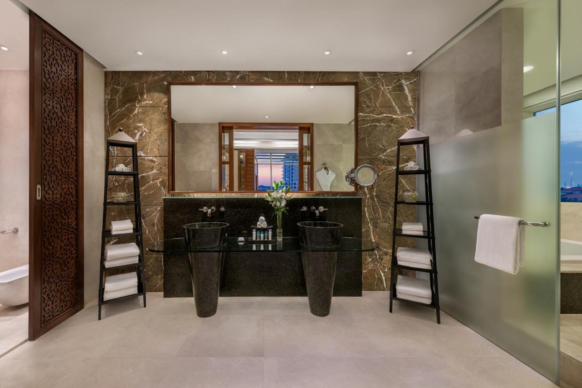 알 라하 비치 호텔 아부다비 외부 사진 The photo shows a modern, elegant bathroom interior. It features a large mirror hanging above a double-sink vanity with a dark stone countertop. On either side of the vanity are tall, slender vases that might be holding decorative elements. There are