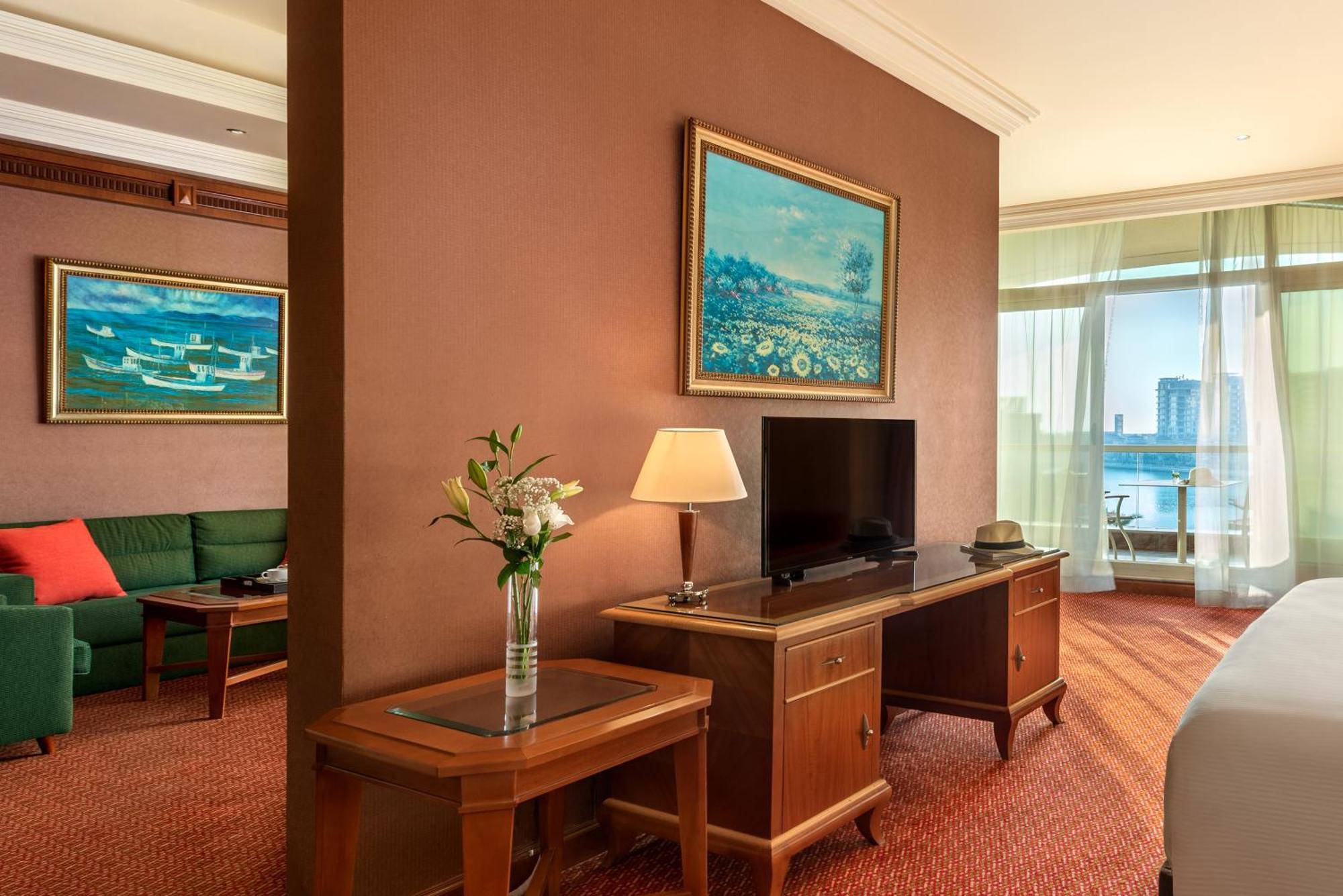 알 라하 비치 호텔 아부다비 객실 사진 The photo shows a hotel room with a warm, inviting atmosphere. There's a large window that provides natural light and a view, likely overlooking a cityscape or water. On one wall, there's a piece of artwork featuring a landscape, and nearby, a small 