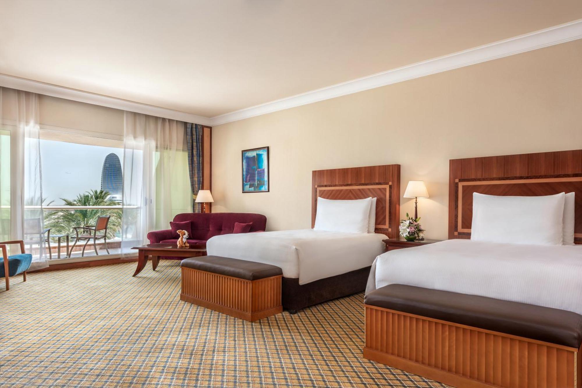 알 라하 비치 호텔 아부다비 객실 사진 The photo shows a hotel room featuring two twin beds with wooden headboards. The room has a light, neutral color scheme, with a large window that provides natural light and a view of the outdoors. There is a small seating area with a purple sofa and 
