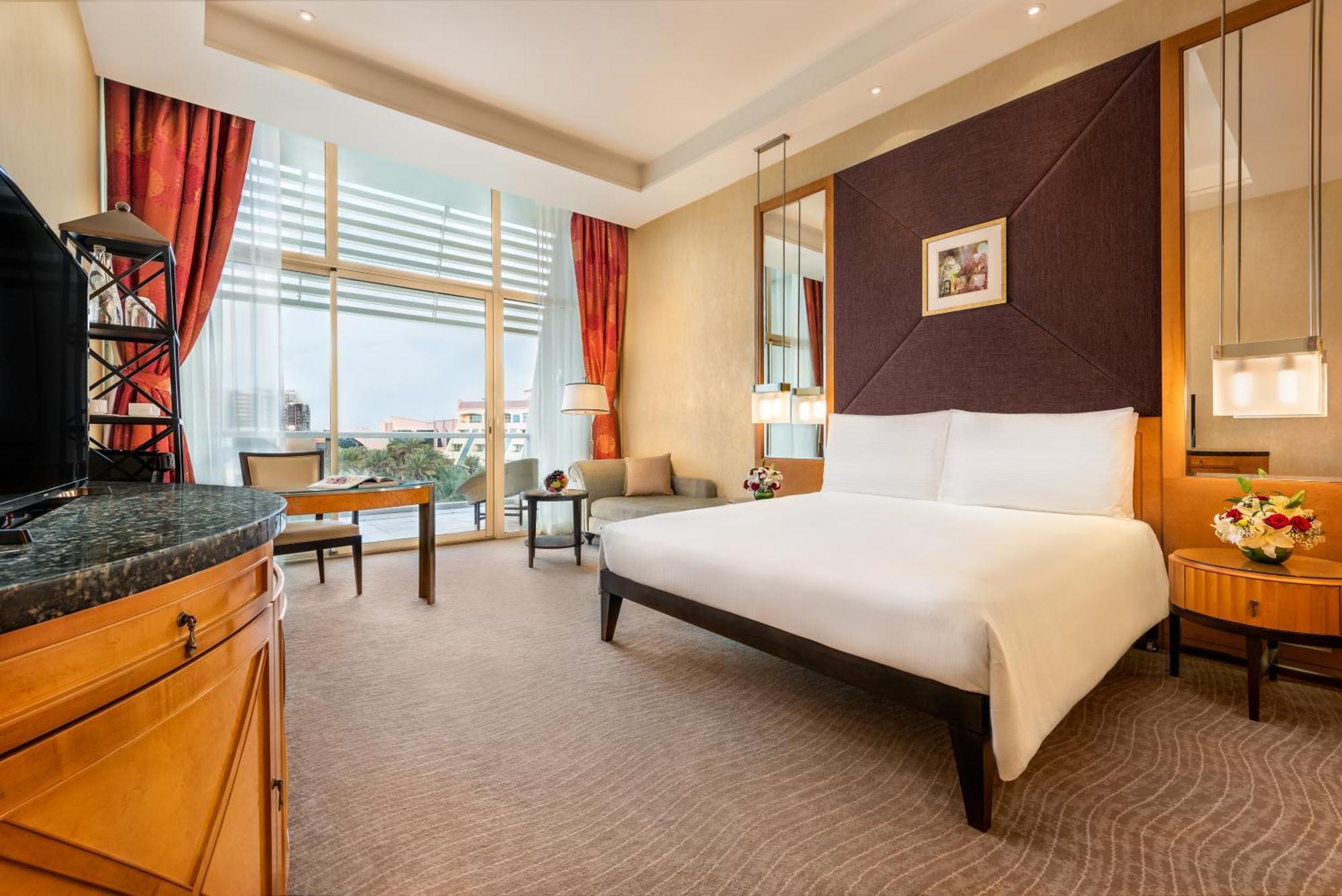 알 라하 비치 호텔 아부다비 객실 사진 The photo shows a modern hotel room featuring a large bed with a white duvet positioned against a purple accent wall. There are tall windows with sheer curtains allowing natural light to enter the space, along with red drapes framing the windows. A d