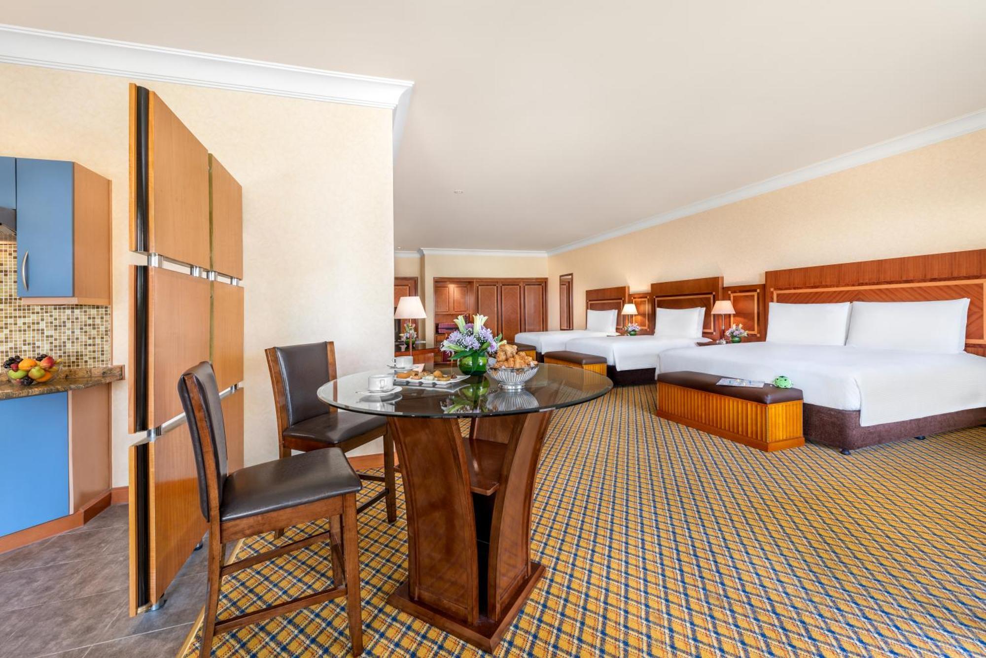 알 라하 비치 호텔 아부다비 객실 사진 The photo displays a spacious hotel room. It features two large beds with white bedding, a circular glass dining table with a decorative floral centerpiece, and a couple of chairs around it. There is a wooden cabinet along one wall, and the room has 