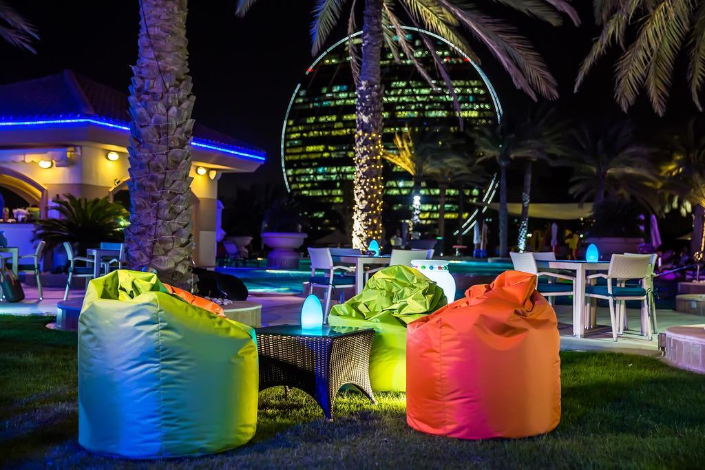 알 라하 비치 호텔 아부다비 외부 사진 The photo shows a vibrant outdoor lounge area at night. In the foreground, there are colorful bean bag chairs—one in bright green, one in yellow, and one in orange. A small table sits between the bean bags. In the background, illuminated palm trees a