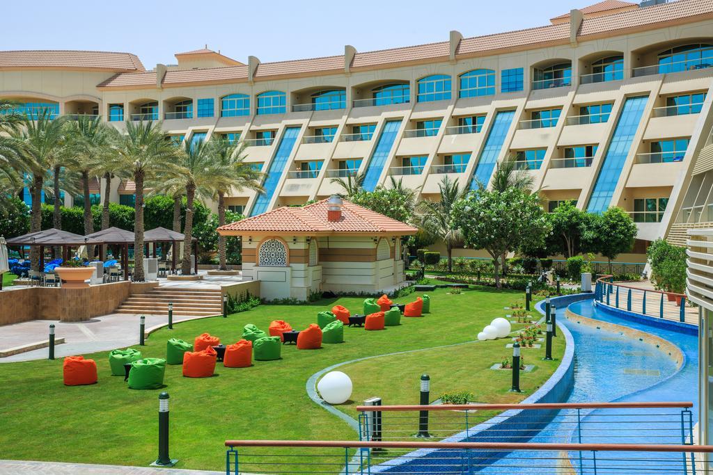 알 라하 비치 호텔 아부다비 외부 사진 The photo depicts a modern hotel complex with multiple stories and large windows. In the foreground, there is a well-maintained lawn featuring colorful lounge chairs in shades of orange and green. A pathway runs alongside a blue water feature, creati
