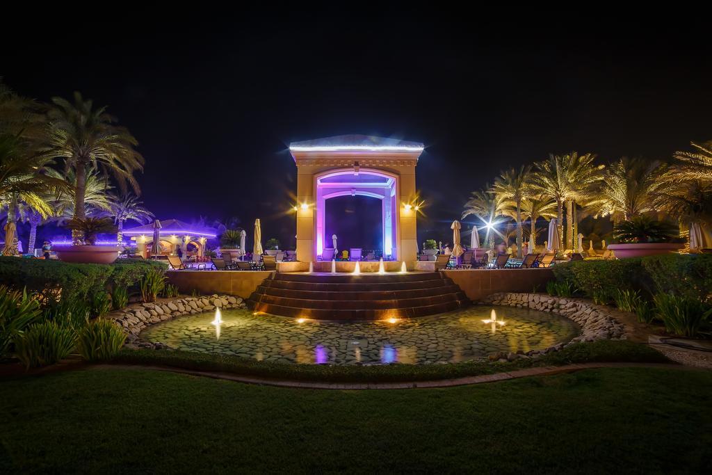 알 라하 비치 호텔 아부다비 외부 사진 The photo depicts a beautifully lit outdoor scene at night. In the foreground, there is a serene water feature with a circular pond, and small fountains creating gentle ripples. Surrounding the pond are lush green lawns and ornamental plants. 

In th