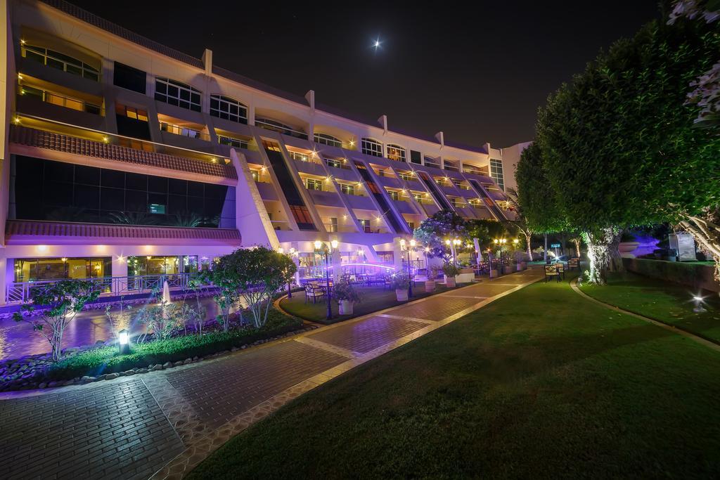 알 라하 비치 호텔 아부다비 외부 사진 The photo shows a modern building illuminated at night, with a vibrant purple hue. The structure has multiple floors, each with balconies overlooking a landscaped area. In the foreground, there is a pathway lined with well-manicured grass and shrubs,