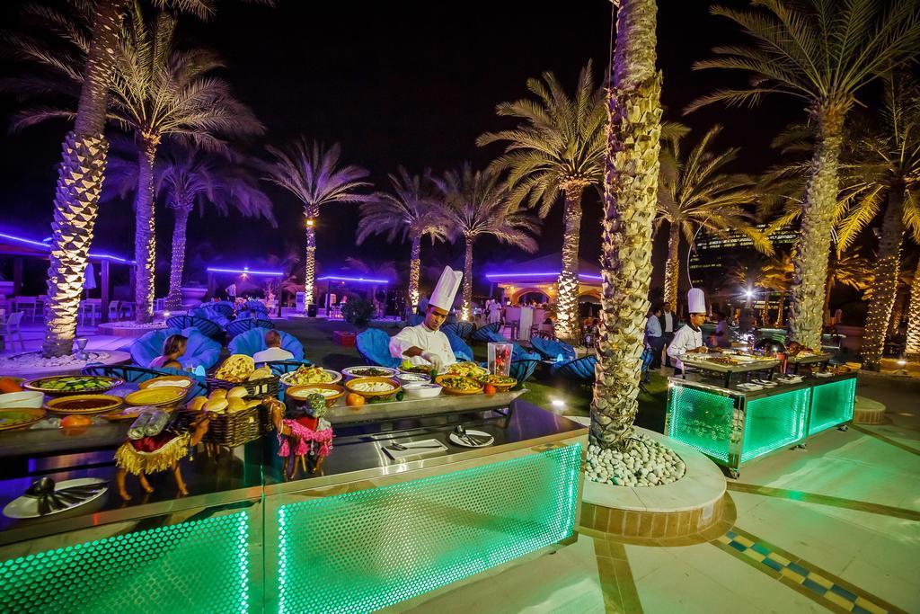 알 라하 비치 호텔 아부다비 외부 사진 The photo depicts an outdoor dining area at night, featuring several chefs in white uniforms and hats working at a food station. The setting is illuminated by colorful lights, with glowing green accents on the serving tables. Surrounding the area are